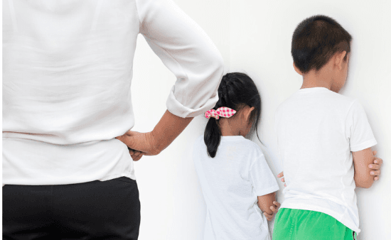 A woman stands with a hand on her hip as a girl and a boy face a white wall with their arms crossed, in timeout.