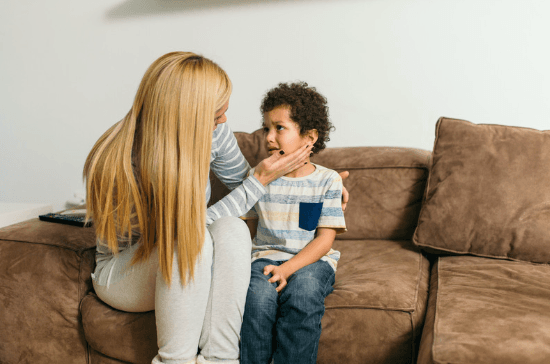 A mother sits on a sofa with her hand on her young son's cheek as he looks up at her, close to tears.
