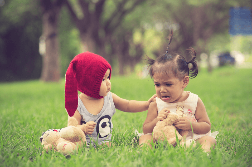 A toddler in a red knit hat reaches out to comfort a crying toddler girl holding a teddy bear on the grass.
