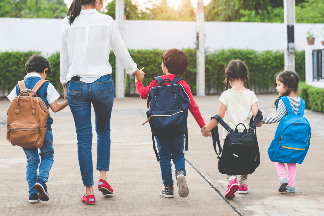 A woman holds hands with a boy as they walk away with three other young children, all carrying backpacks, toward school.