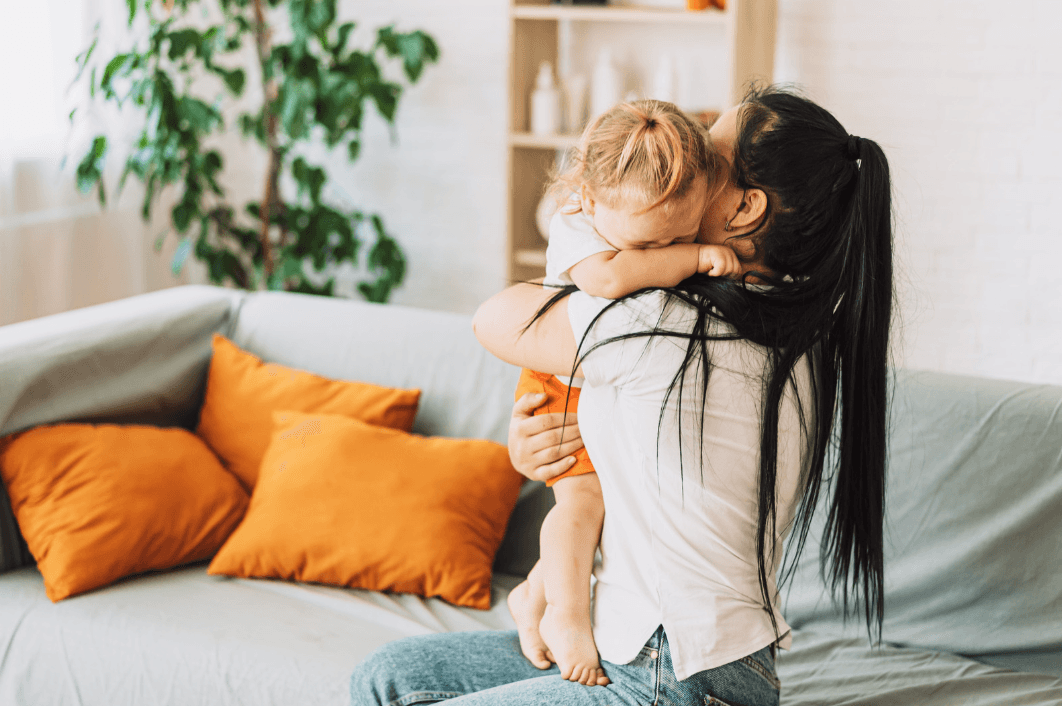 A mother in a white shirt holds her small child, who buries their face in her shoulder, on a sofa with orange cushions.