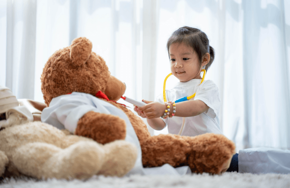 A young girl in a toy stethoscope plays doctor with a large teddy bear in a white coat.