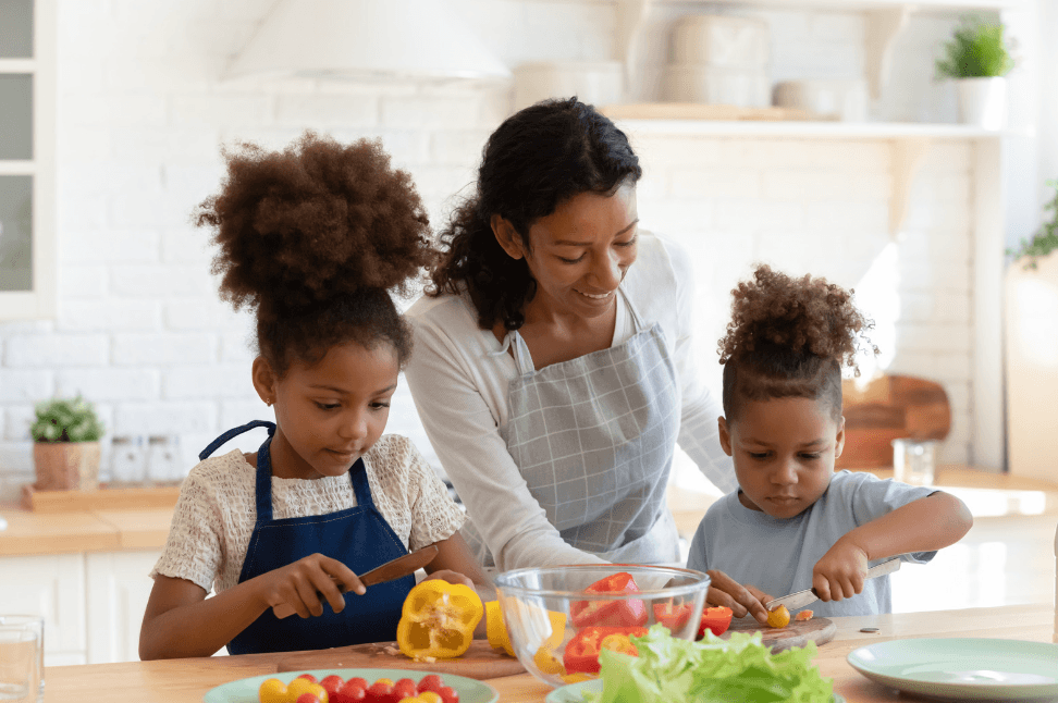 A mother helps her daughter and young son chop peppers and tomatoes together in a bright kitchen.