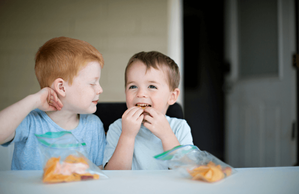 Two young brothers sit at a table with bags of snacks, one eating happily while the other looks at him.