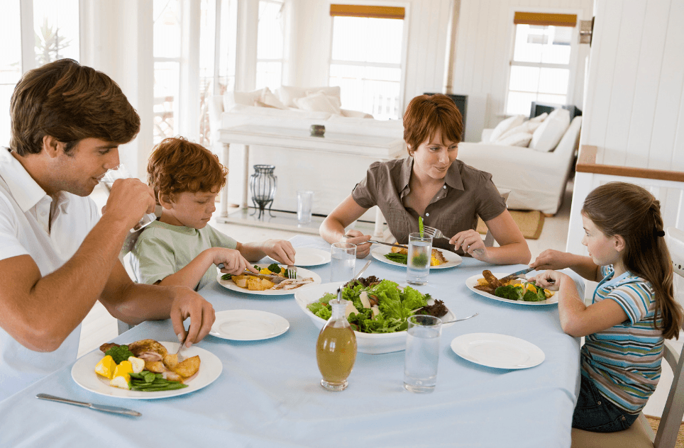 A family of four eats a meal together at a set dining table with salad, water and plates of food.