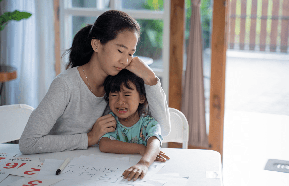 A mother rests her cheek on her crying young daughter's head at a table with number flashcards and homework.