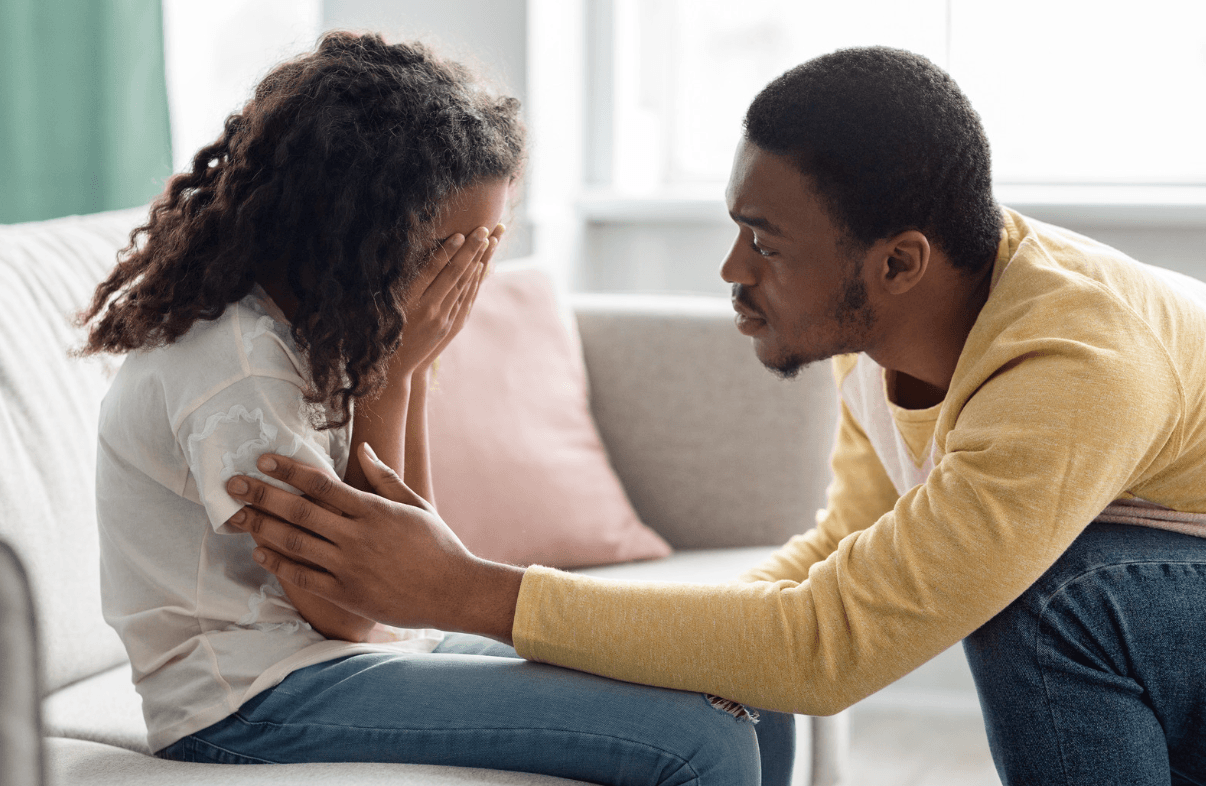 A father leans toward his daughter, who sits on a sofa with her hands over her face, and gently touches her arm.