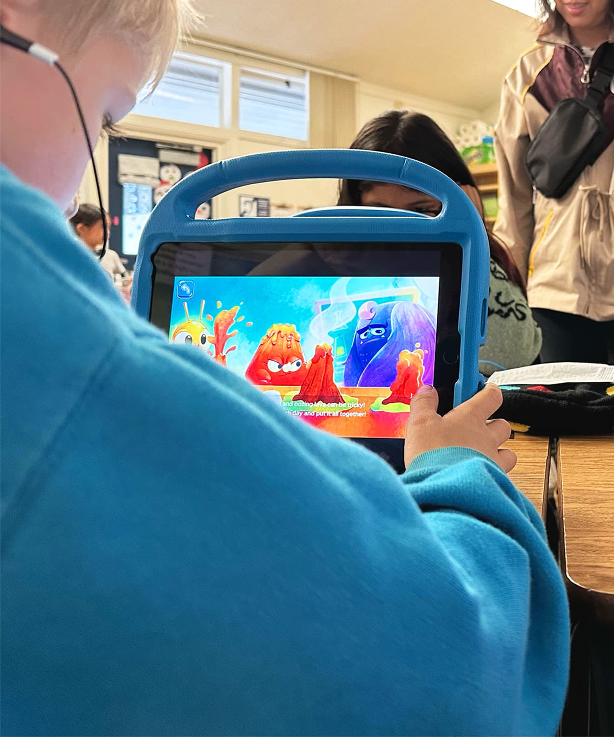 A child in a teal sweater holds a blue-cased tablet showing MEandMine emotion buddies in a classroom.