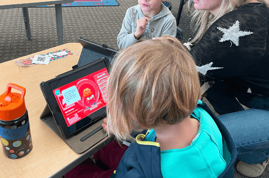 A child at a classroom table views the Dinomite emotion buddy profile on a tablet while an adult sits beside them.