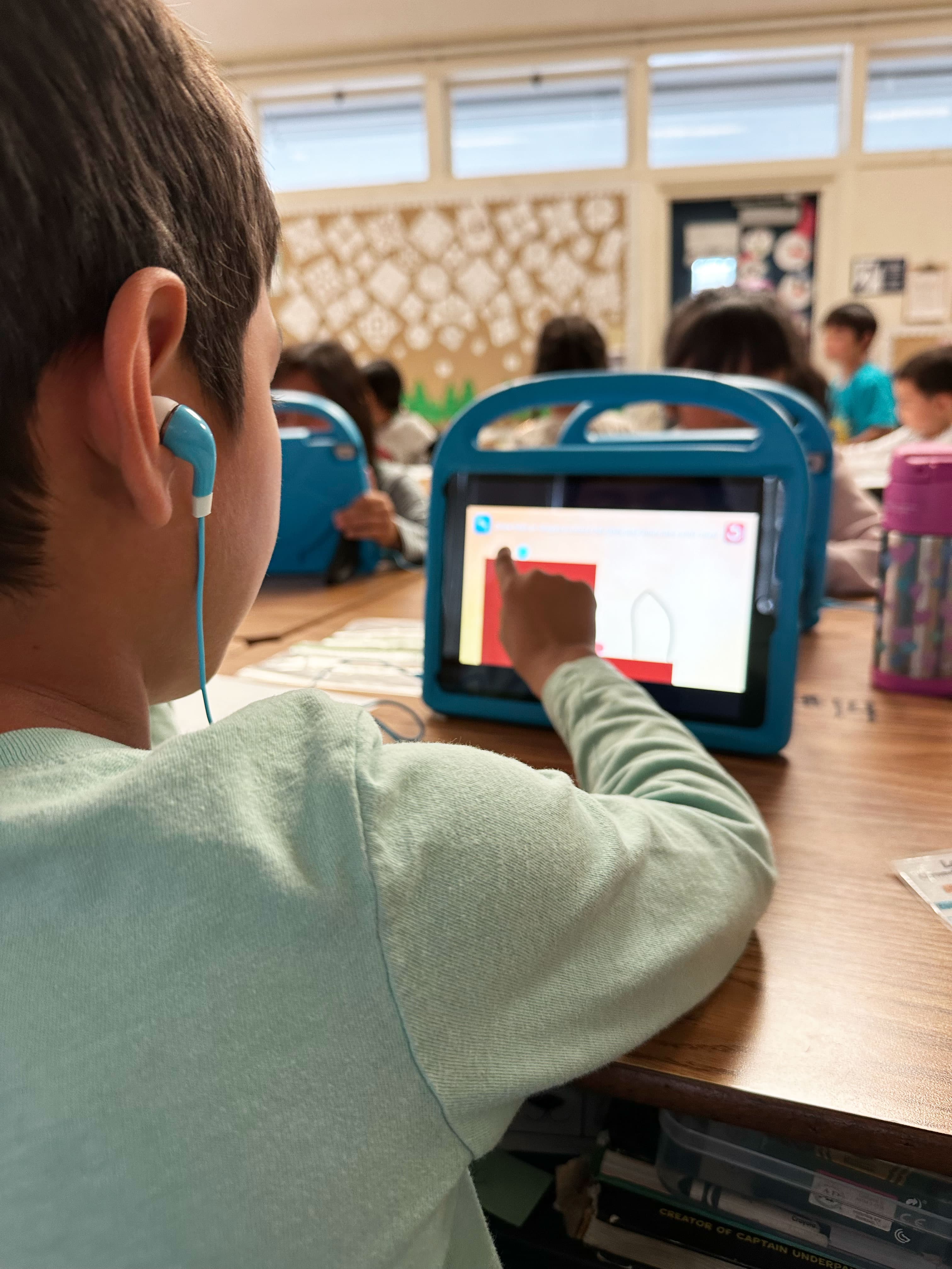 A boy wearing earbuds traces a shape in a MEandMine game on a blue-cased tablet in a busy classroom.