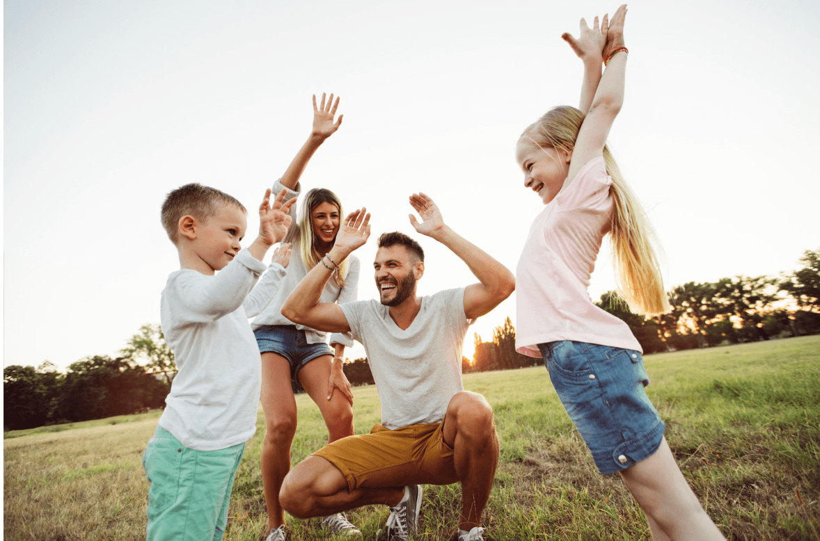 A family of four laughs with hands raised in a sunny field, cheering each other on