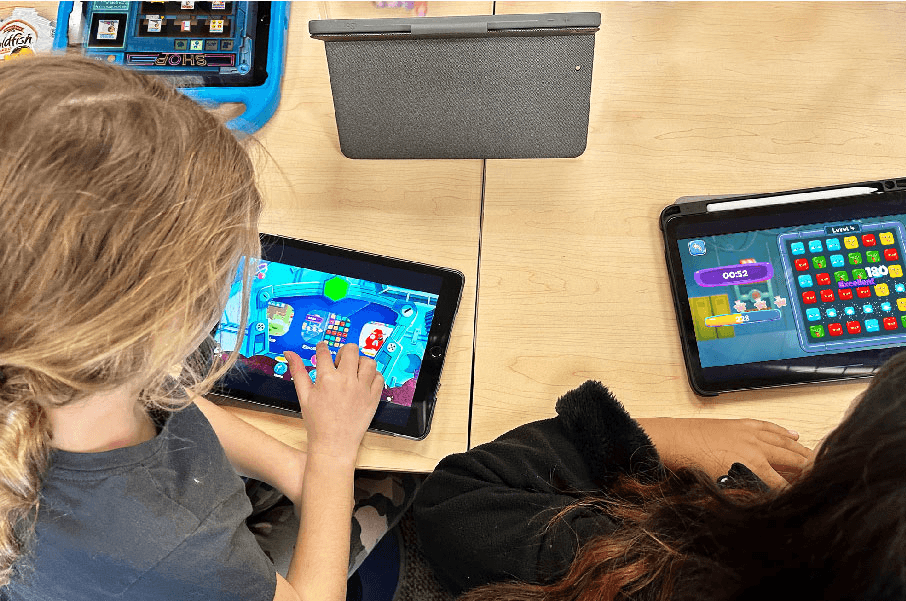 Two students play MEandMine games on tablets at a classroom table, one on a tile-matching level.