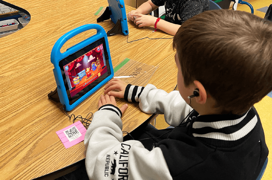 A boy wearing earbuds plays a MEandMine game on a tablet in a classroom, with a stage of musical characters on screen.