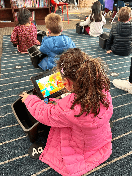 A girl in a pink jacket sits on a classroom carpet watching a MEandMine game on a tablet while other children sit nearby.