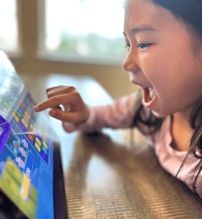 A girl laughs as she taps a MEandMine game on a tablet at a table.