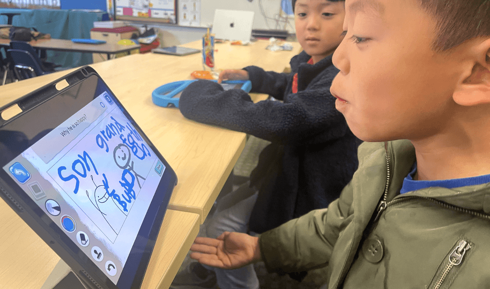 A boy in an olive jacket speaks beside a tablet showing a stick-figure drawing, as a classmate looks on.
