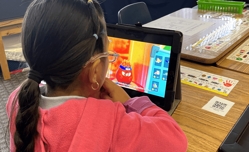 A girl in a pink sweatshirt watches a MEandMine game on a tablet where a red emotion buddy earns rewards.