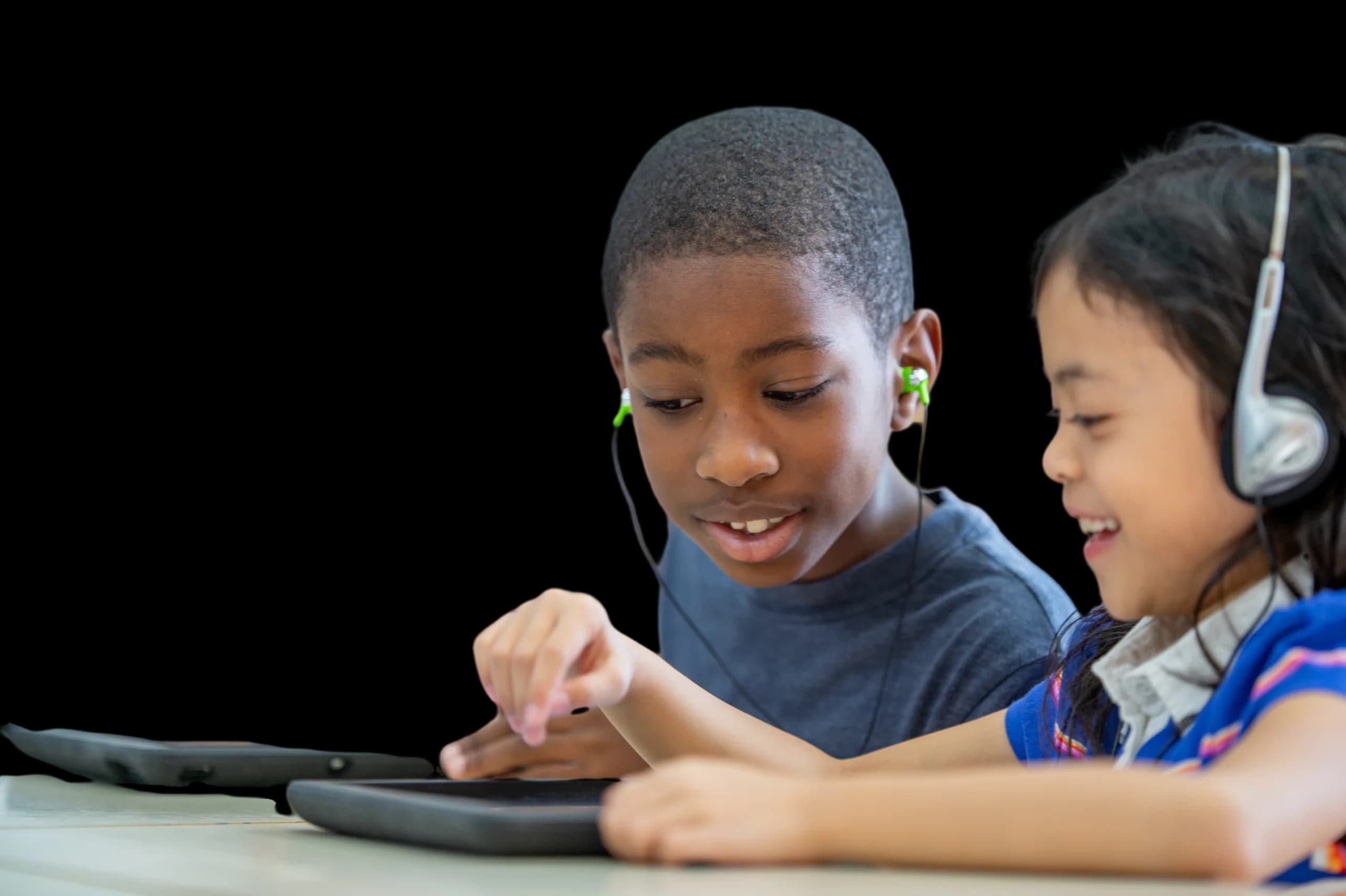 Two students smiling as they play on a tablet together, wearing headphones