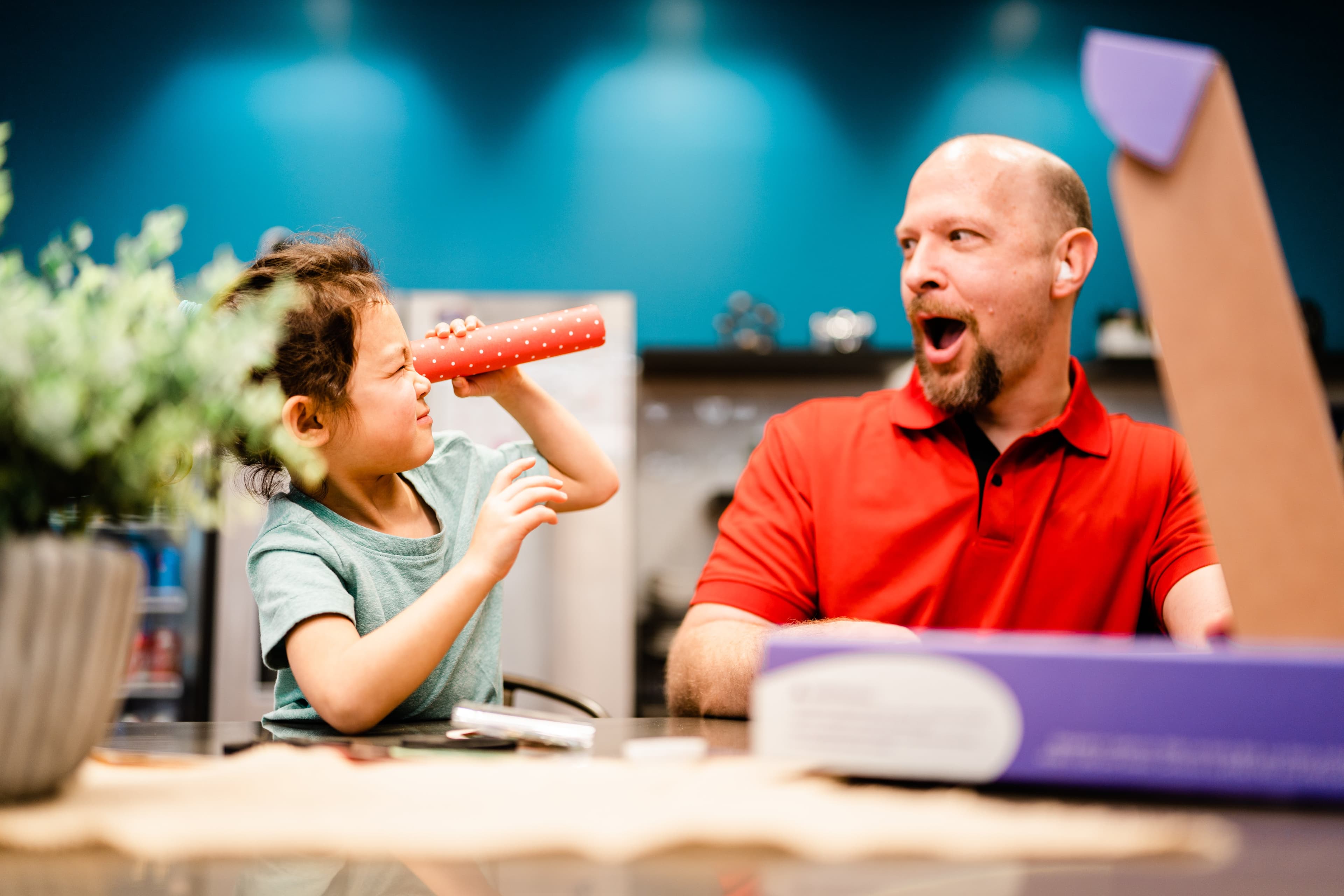 A child peers through a red polka-dot tube like a telescope while a man in a red shirt reacts with surprise