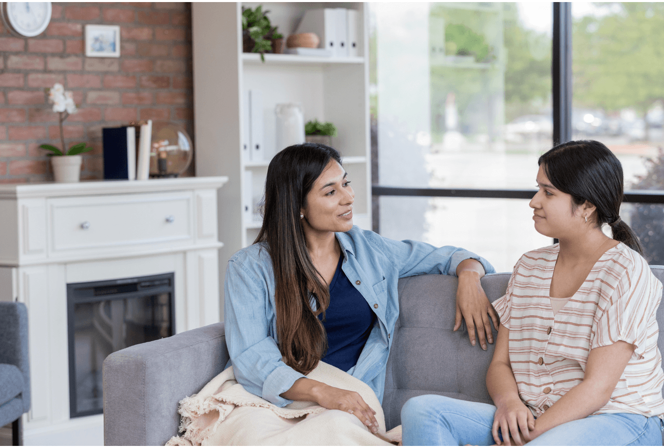 A mother sits on a sofa talking warmly with her teenage daughter, who looks back at her