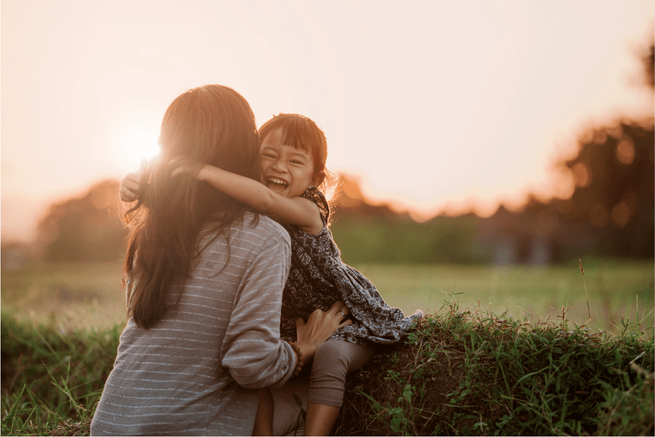 A laughing little girl hugs her mother in a grassy field at sunset
