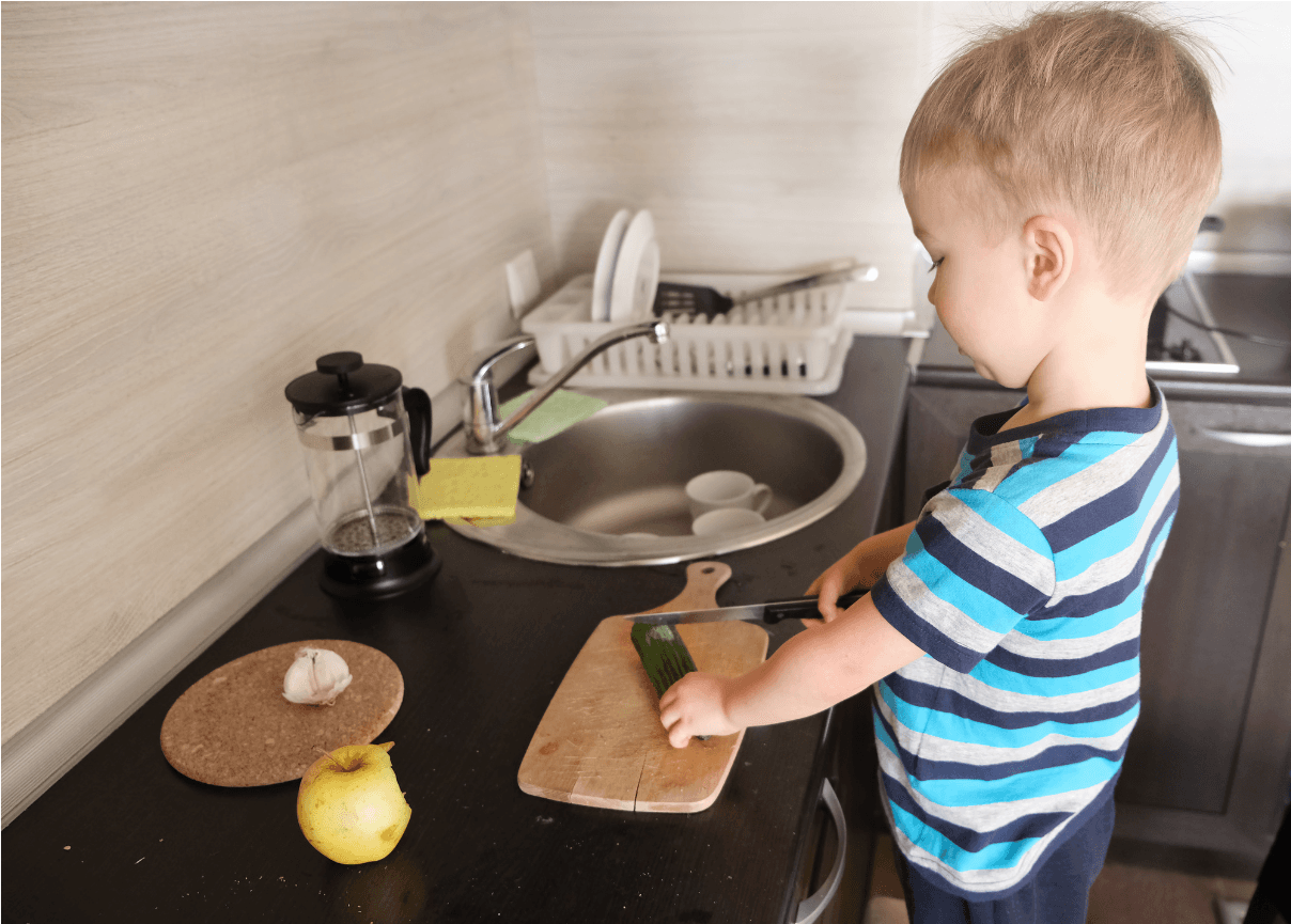 A toddler in a striped shirt stands at a kitchen counter carefully cutting a cucumber on a board