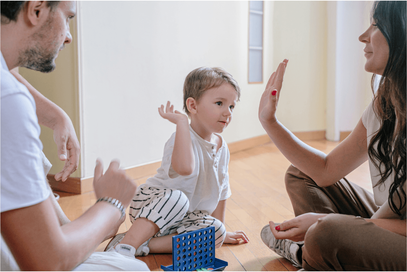A toddler raises a hand to high-five his mother while his father looks on, with a Connect Four game on the floor