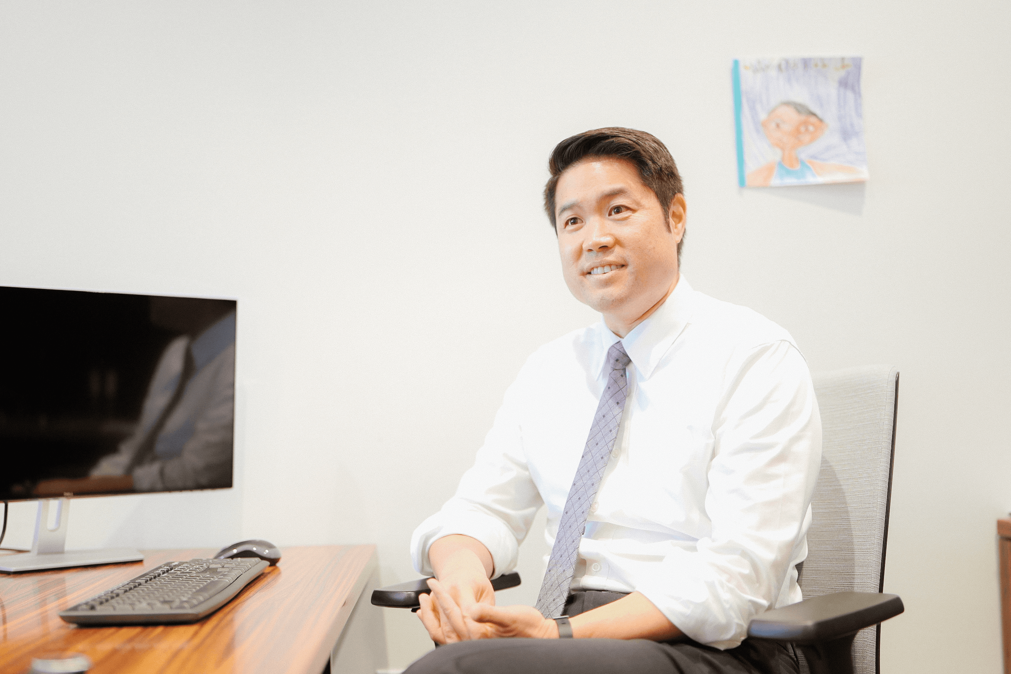 Smiling pediatrician in a white shirt and tie seated at a desk in an office, with a child's drawing on the wall