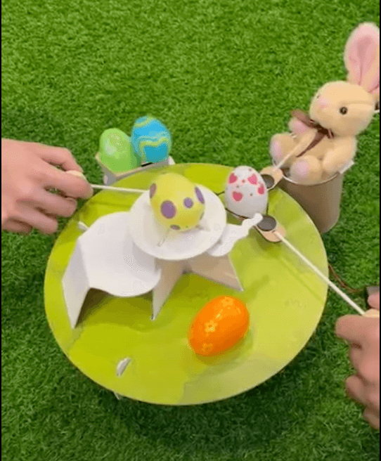 Hands use sticks to move painted Easter eggs on a round green platform, with a plush bunny and extra eggs nearby.