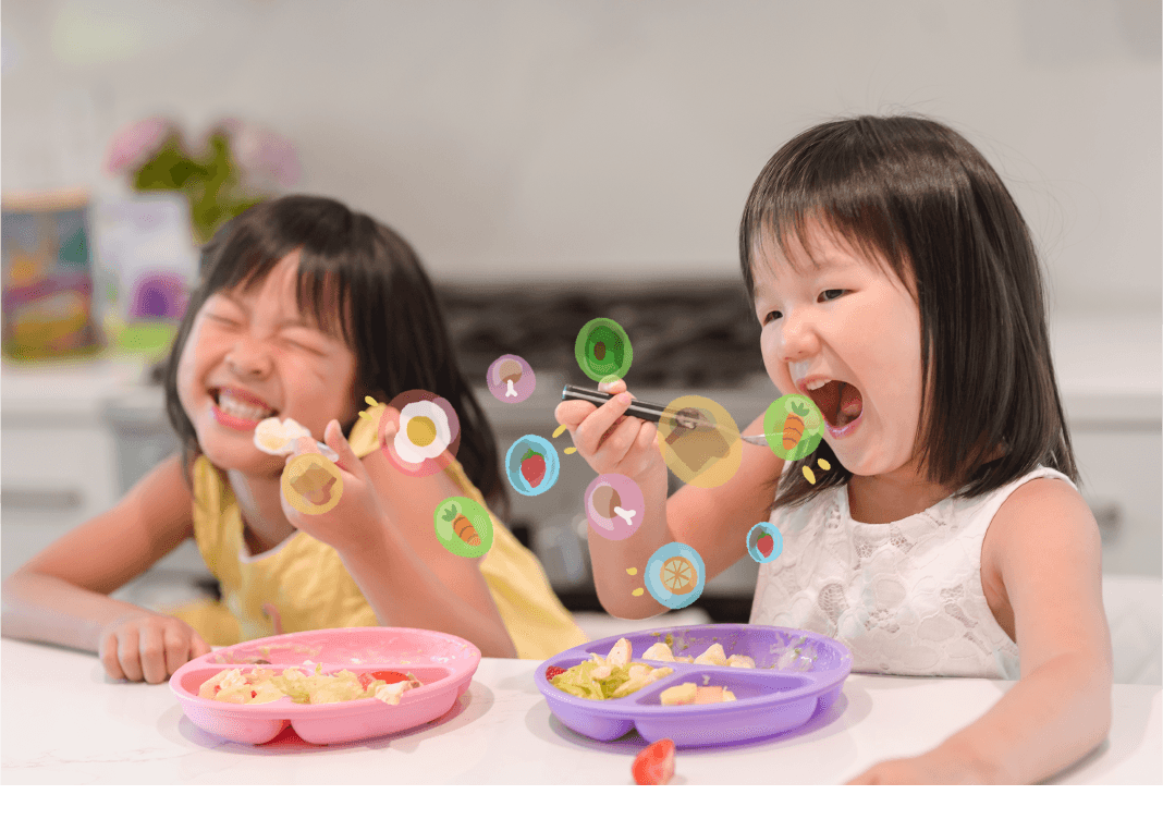 Two laughing girls eat from pink and purple plates, with colorful food icons like carrots and eggs floating around them.