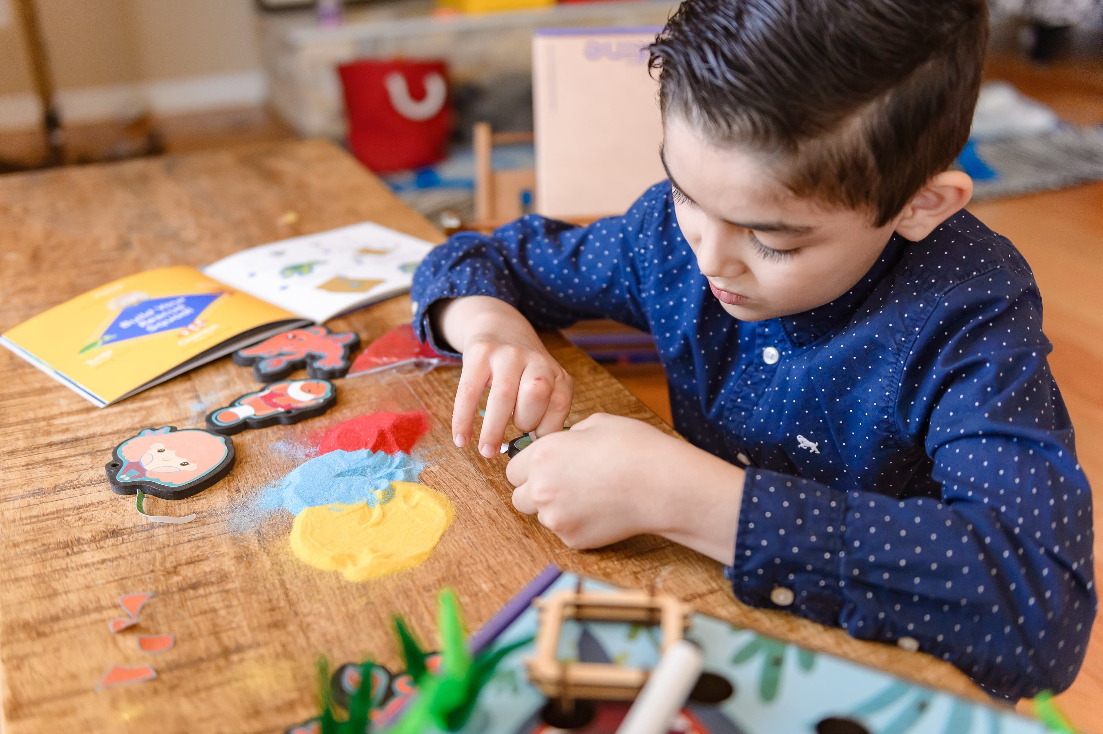 A boy in a blue dotted shirt concentrates on a craft at a wooden table with colored sand, figures and an activity booklet.