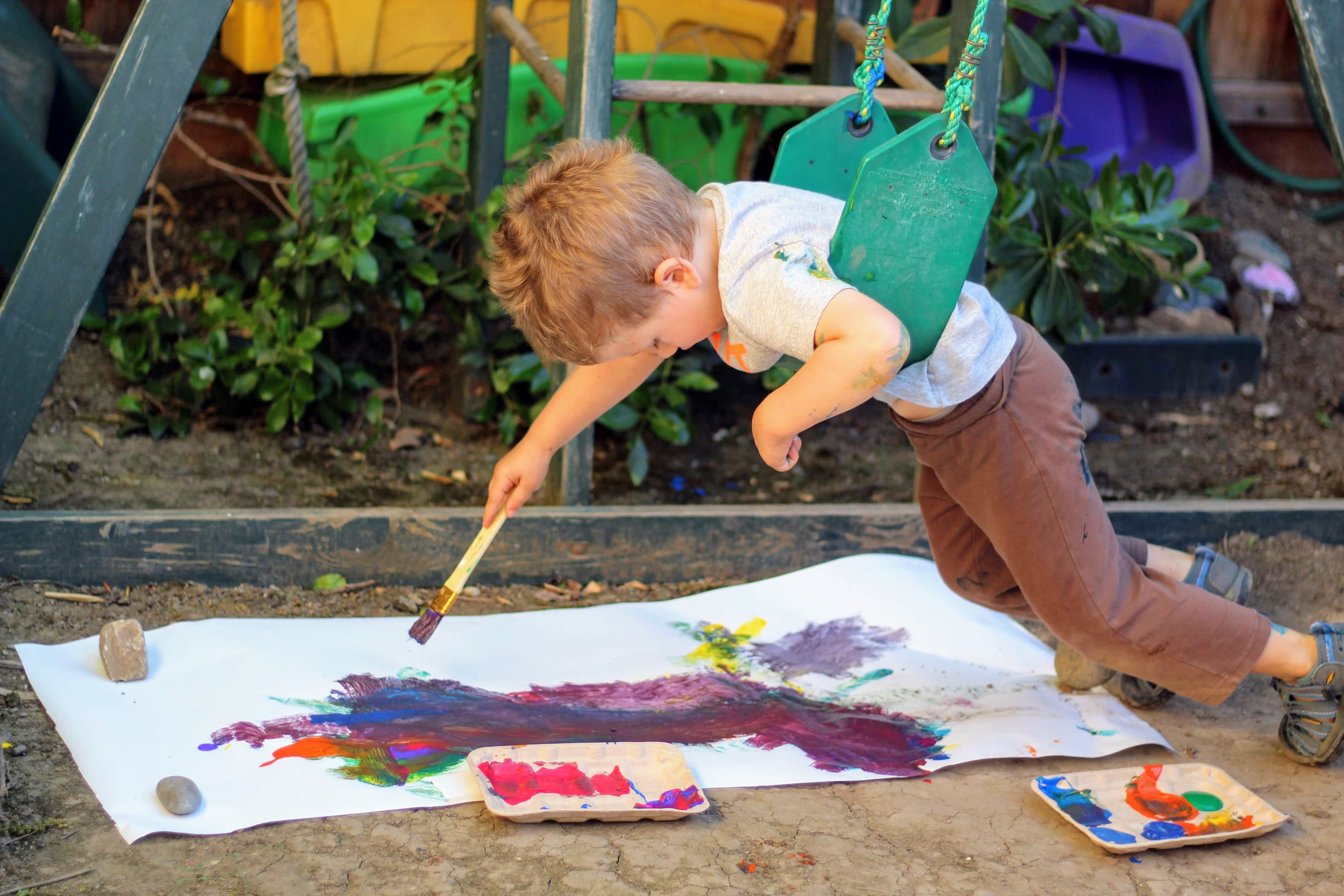 A young boy leaning from a swing to paint with a brush on a long sheet of paper outdoors, with paint trays beside it