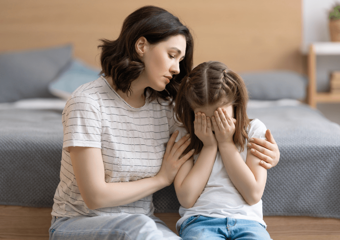 A mother sits on the floor beside a bed with her arm around her young daughter, who is crying into her hands.
