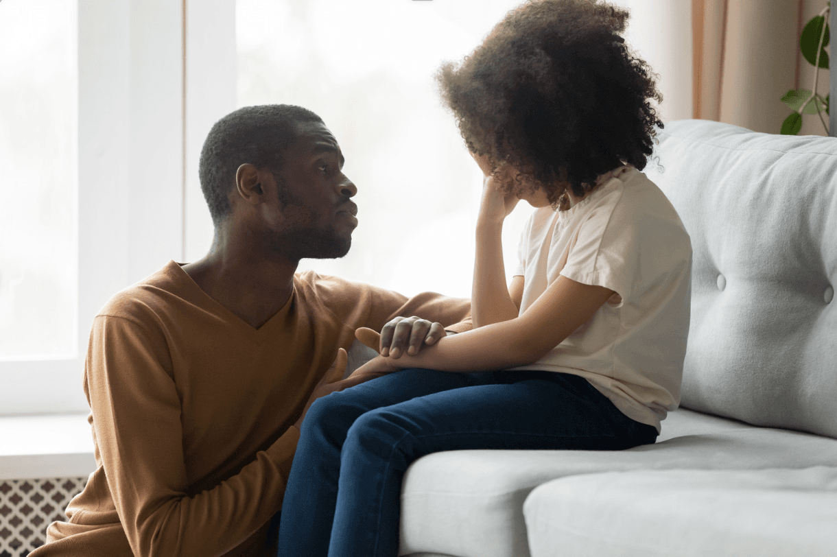 A father kneels beside his upset daughter on a sofa, holding her arm and speaking to her gently.