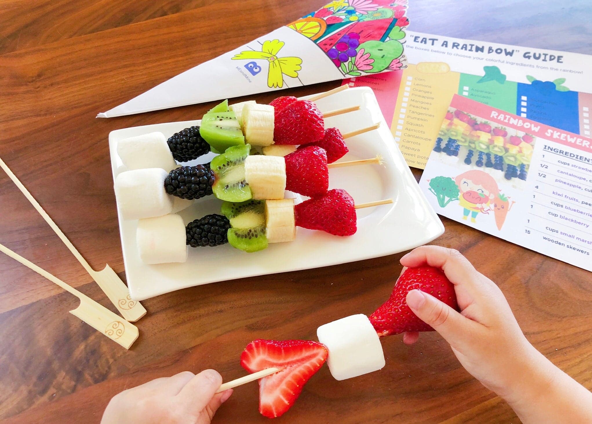 A child's hands assembling a fruit skewer beside a plate of rainbow skewers, a bouquet cone and a recipe guide