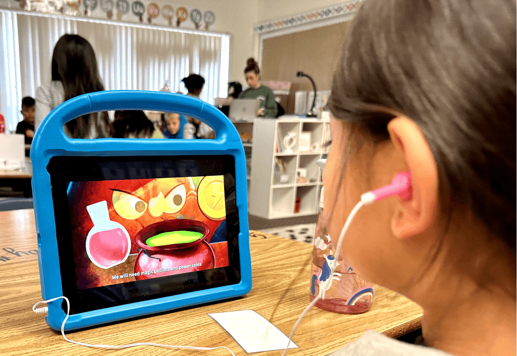 A girl wearing earbuds watches a MEandMine story on a tablet in a classroom, showing a potion pot and an angry character.
