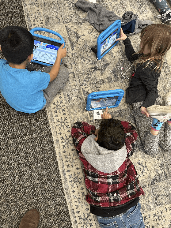Three children lie and sit on a classroom rug using blue-cased tablets to play MEandMine.