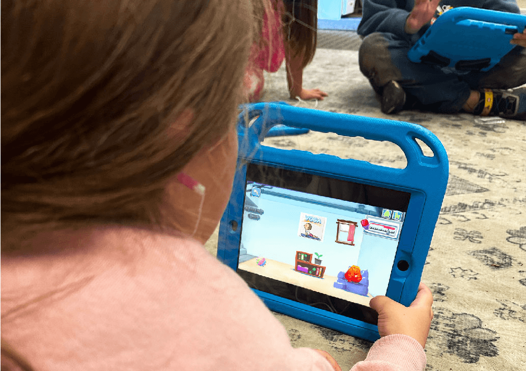 A girl with earbuds holds a blue-cased tablet showing a calming corner room with a yoga card and a red emotion buddy.