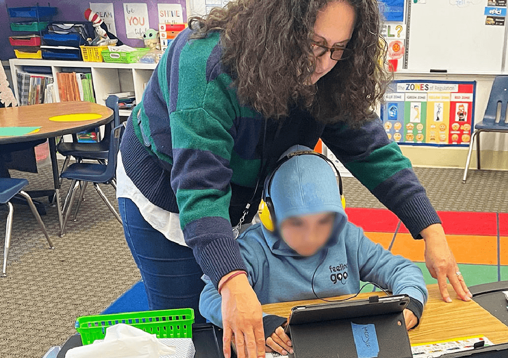 A teacher leans over a boy in a hoodie and headphones using a tablet, with a Zones of Regulation poster behind them.