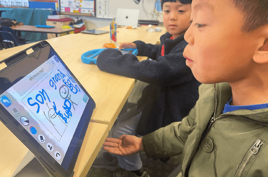 A boy in an olive jacket explains a tablet drawing of stick figures and words while a classmate watches in class.