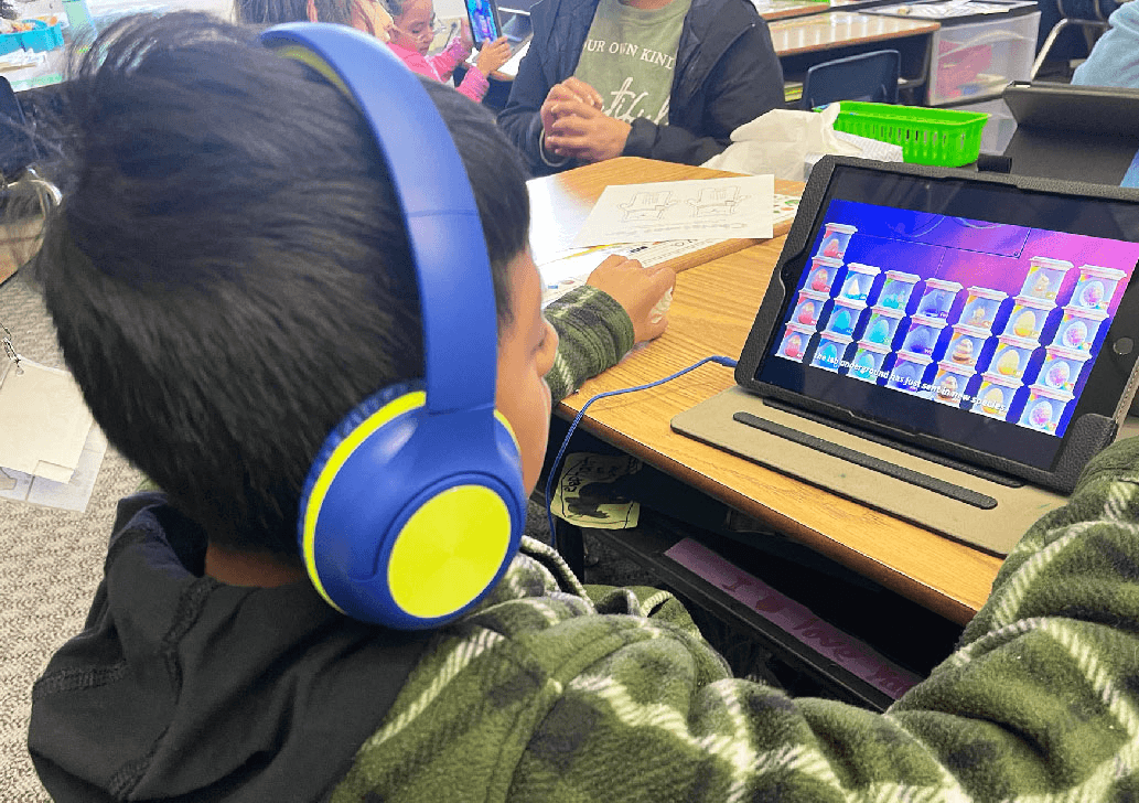 A boy in blue headphones watches a MEandMine game on a tablet showing rows of colorful emotion buddy eggs.