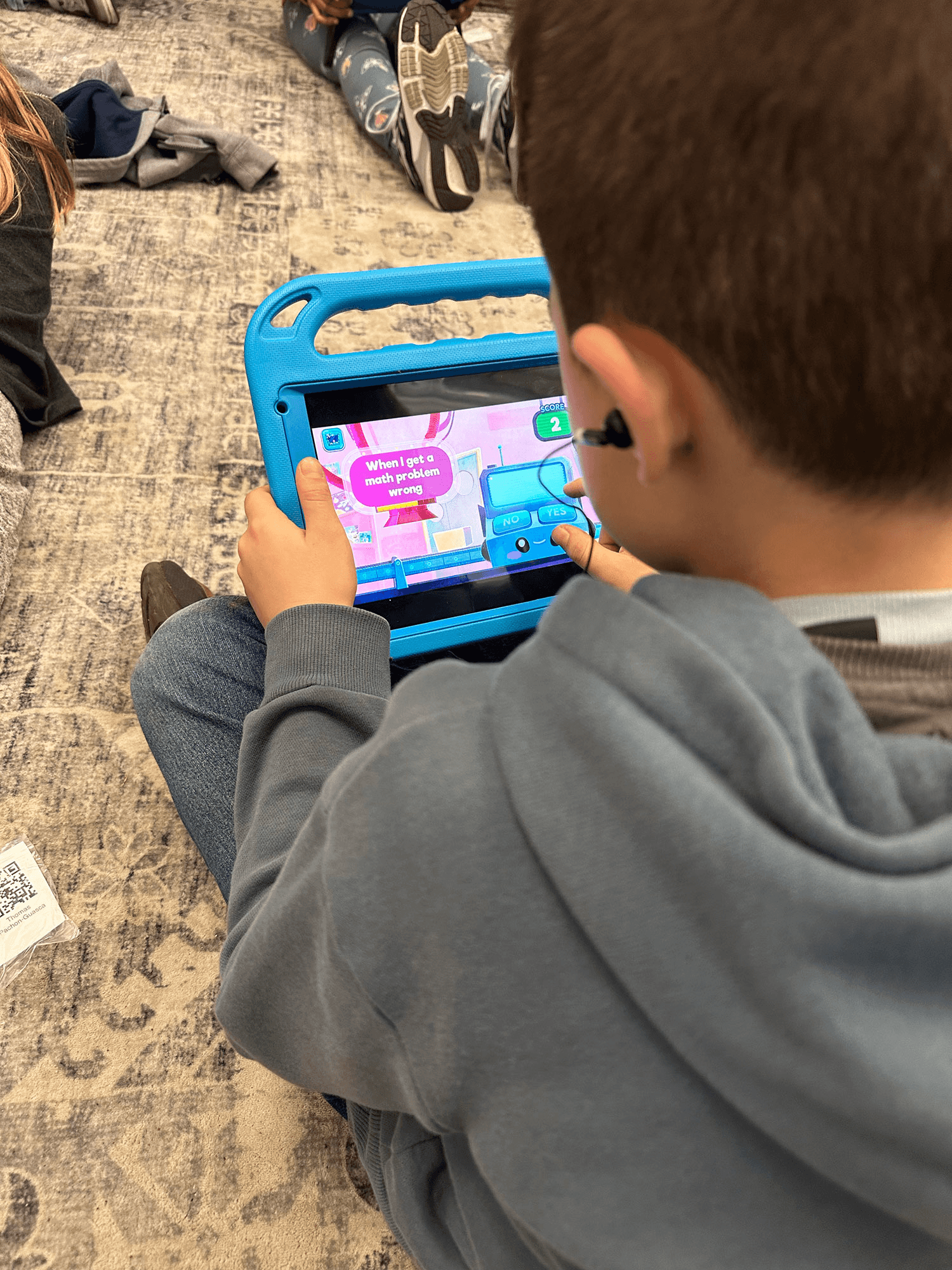 A boy with an earbud on a classroom rug taps yes or no on a MEandMine tablet game asking about a math problem going wrong.