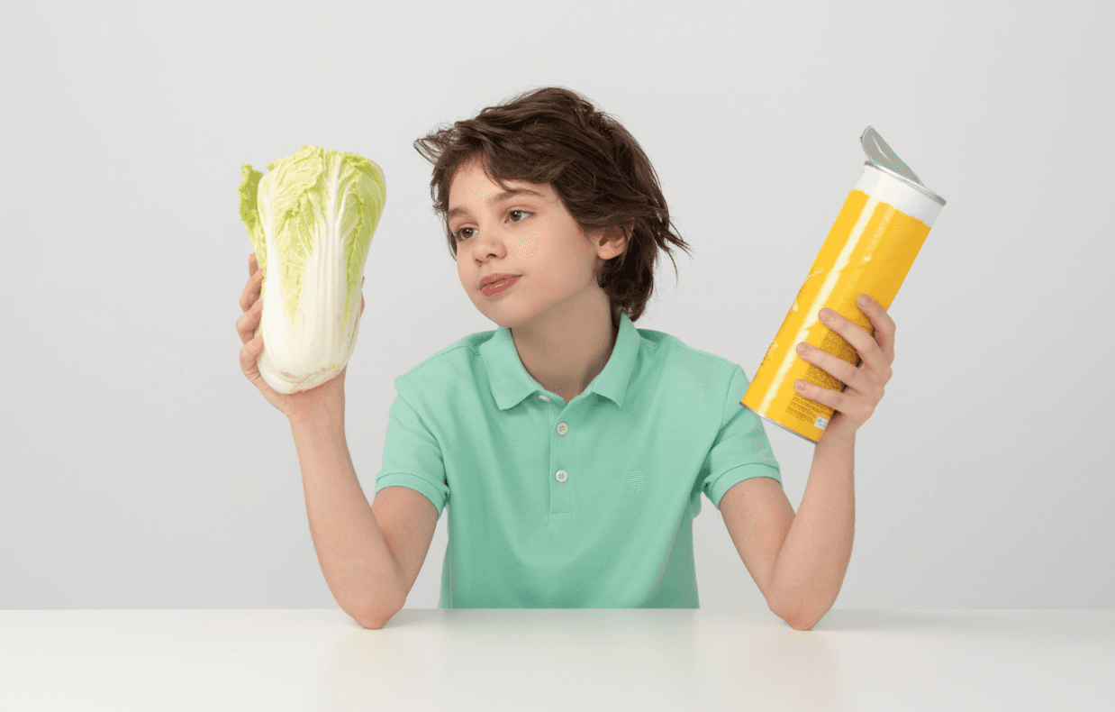 A boy weighs a head of cabbage in one hand against a tube of chips in the other, deciding between them