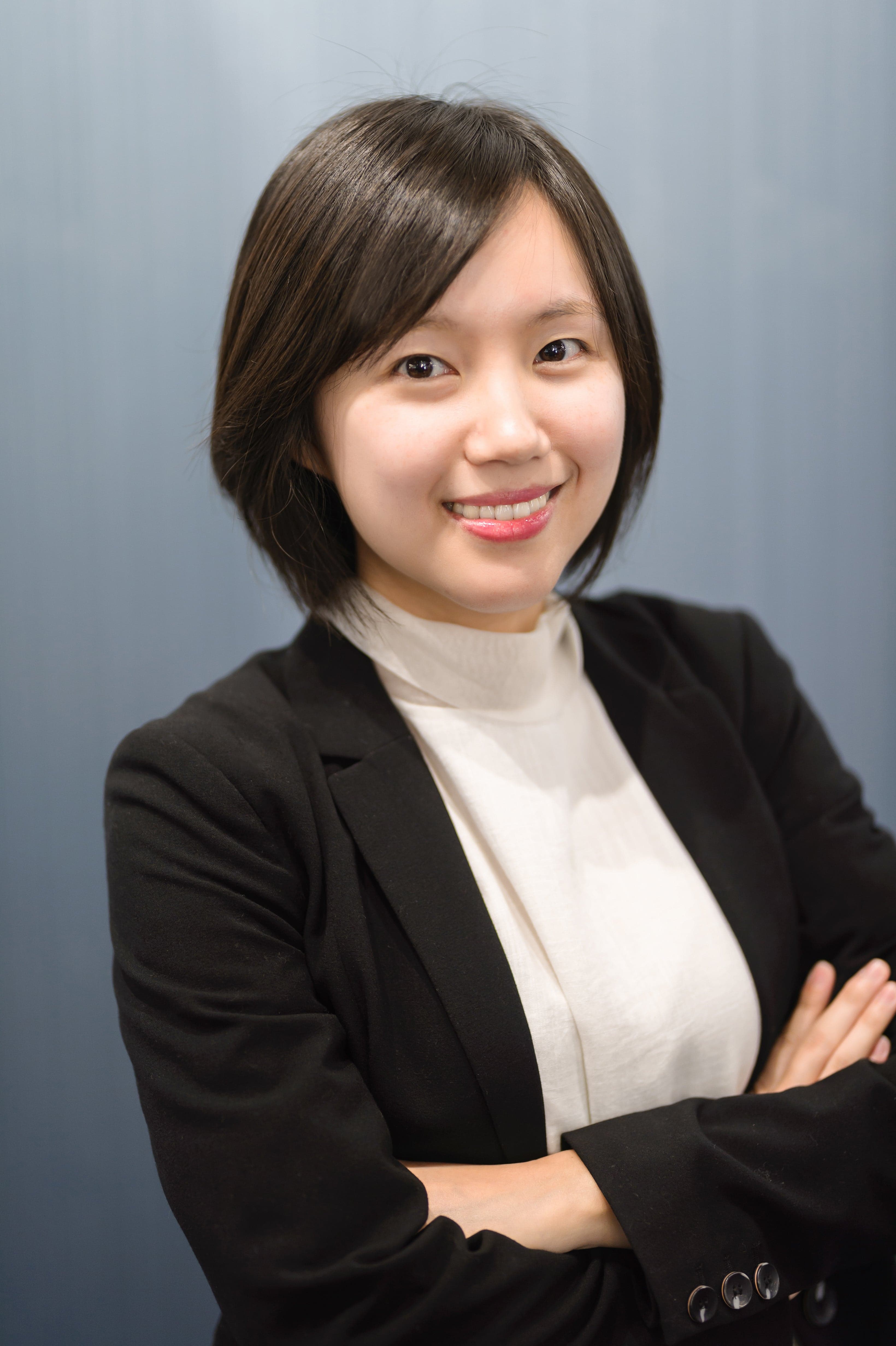 Portrait of a smiling woman with short dark hair in a black blazer and white top, arms crossed, against a blue-gray backdrop