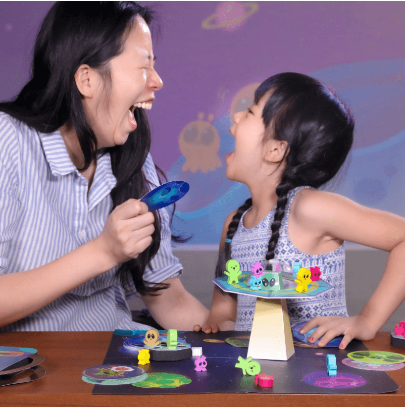 A laughing mother and daughter play the MEandMine iCalm Galaxy board game, with alien figures on a flying saucer piece.