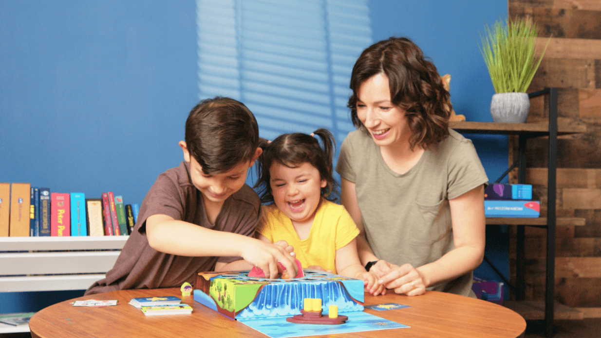 A mother and two laughing children play a colorful waterfall-themed board game together at a round table