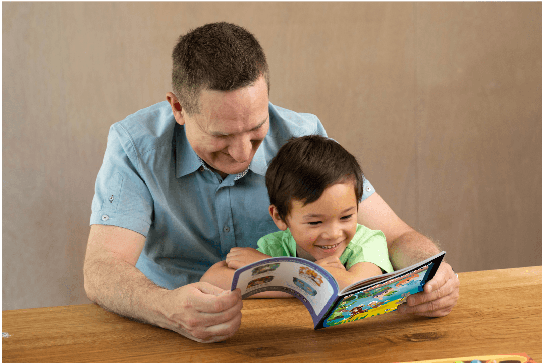 A father and his smiling young son read an illustrated activity book together at a wooden table